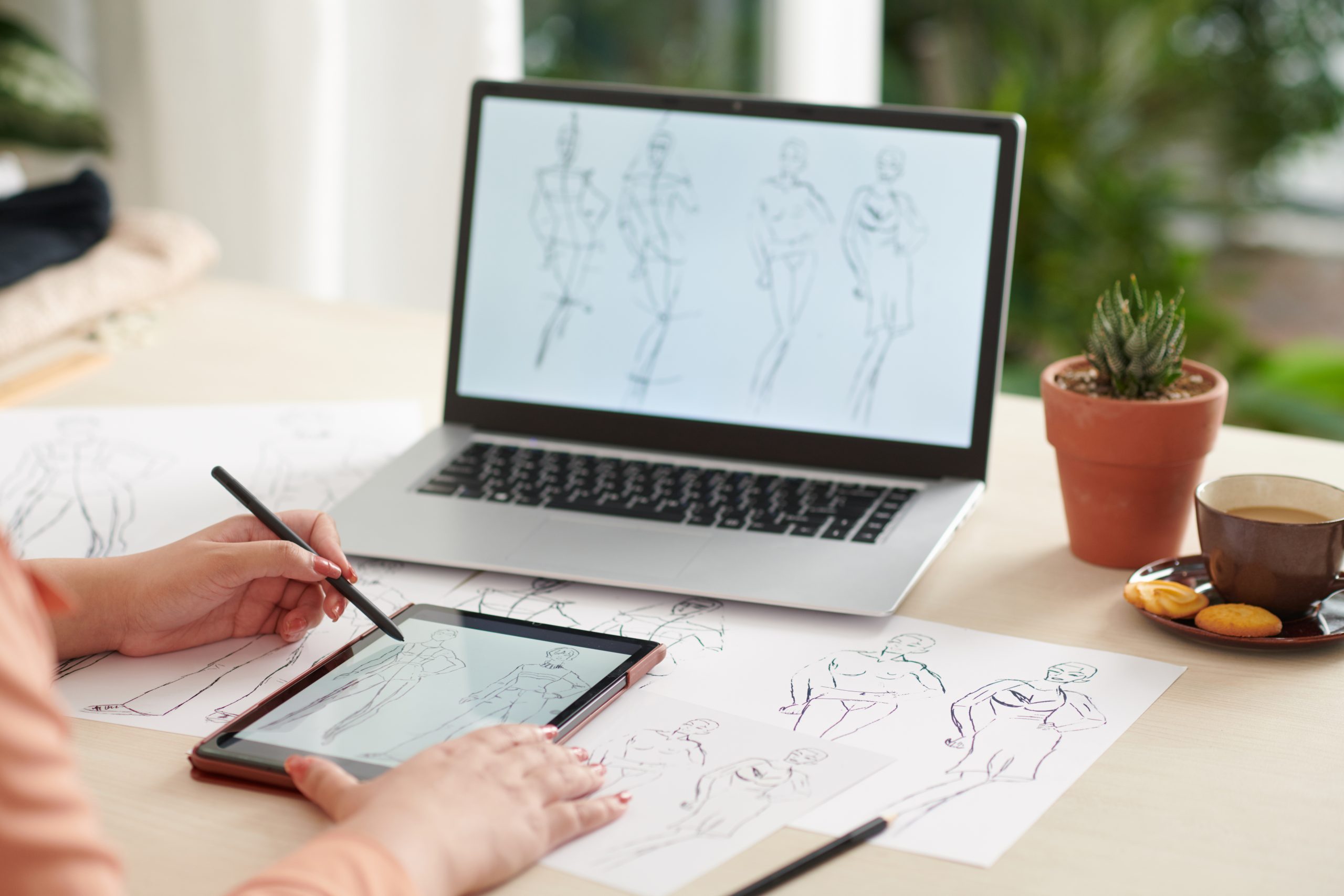 Persona dibujando bocetos de moda en una tableta digital, con ilustraciones de figuras humanas en papel y en la pantalla de una laptop.
